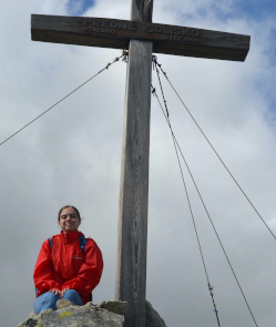 A young woman sitting on a rock posing with a large wooden cross found at the peak of a mountain in the High Tatras