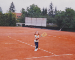 Young girl with a tennis racket in her hands standing on a clay tennis court with trees in the background